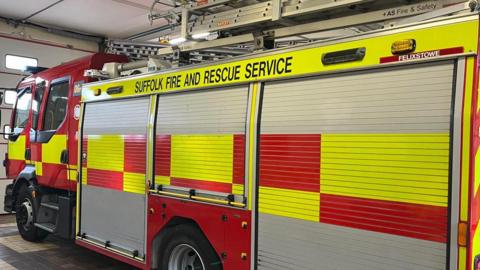 A fire engine, in a fire station, with doors shut. It has ladders on the top, and yellow and red signage. 