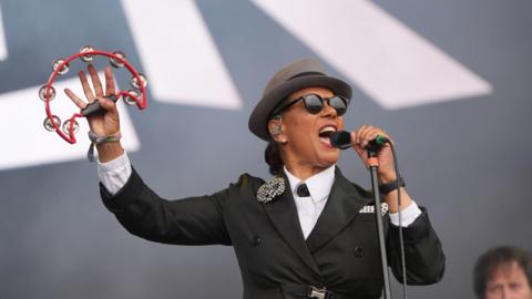 Pauline Black of The Selecter performing on stage while holding a tambourine. She wore all black, black sunglasses and a hat. 