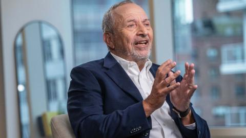 Larry Summers is talking to someone out of shot. He is cropped at the waist. He is wearing a  blue suit and he is using his hands to express his point