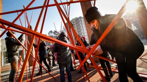 People are using lots of brightly coloured wooden sticks to create structures in a town square