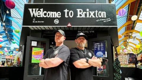 Pete and Daniel are stood almost back-to-back with crossed arms, but facing towards the camera, with straight faces. They are stood below their shop with the wording 'Welcome to Brixton' in black and white on the front.  They are wearing branded black T-shirts and black caps. 