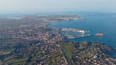 An aerial shot of Guernsey showing a harbour and a bay with boats, buildings, green spaces and trees with a blue sky.