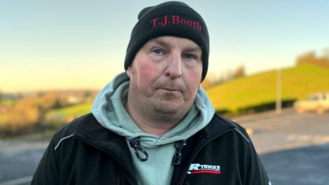 Jonathan Potter is looking directly at the camera. He has pursed lips and is wearing a black beanie hat, a grey hoodie and a black jacket. There are green fields in the background.