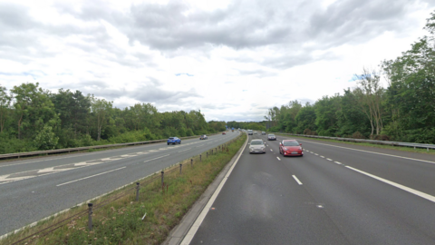 The motorway has six lanes with a grass strip and a small fence down the middle. There are about a dozen cars on the rod. The road is bordered by tall green trees.