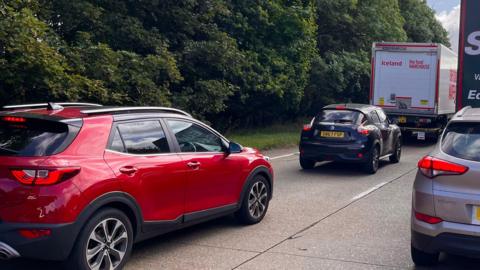 Two lanes of queuing traffic on a dual carriageway. Two large lorries can be seen and three cars. 