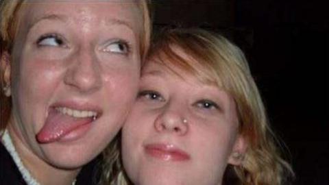 A close-up image of two young women looking into the camera. The woman on the left is looking off to the side and sticking her tongue out. The woman on the right is looking into the camera. 