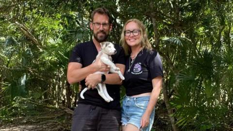 David Brudnell is holding a small light-coloured puppy in his arms. He has a dark T-shirt on and has short dark hair and a beard. He is standing next to Kathryn Grace who has long blonde hair and is wearing a T-shirt and shorts. There are trees behind them. 