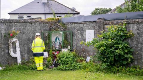 A worker in yellow high visibility clothing at a memorial left at the site of the former Mother and Baby Institution in Tuam, County Galway.