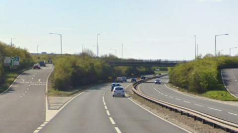 The A47 at the Saddlebow roundabout near King's Lynn. Slip roads up to the roundabout, which is on higher ground, can be seen. There are cars driving away from the camera travelling westbound.