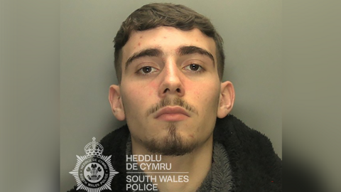 A custody image of a man looking at the camera. He has dark brown hair and a dark stubbly beard and moustache.