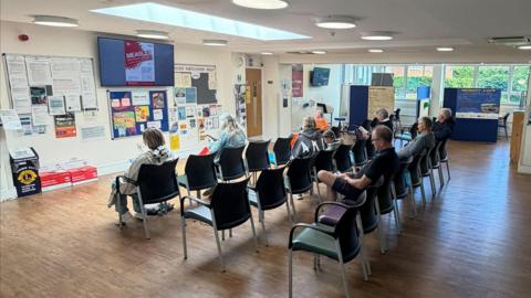 An image of the reception area at Oxted Health Centre. There are several rows of chairs with people sat in them.