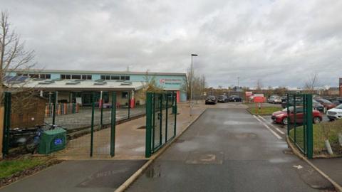 The outside of Bourne Elsea Park Primary Academy. It shows a road leading to a car park. There are green gates which are open. The sky is overcast.