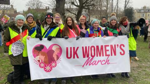 A group of women, mostly wearing hi-vis jackets, hold a banner saying UK Women's March, which includes a graphic of a heart featuring four women and flowers. The group is standing on a patch of grass with trees behind.