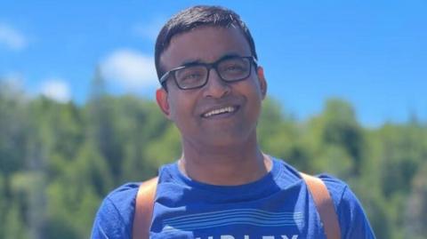 Sharif Rahman smiling straight at the camera. He is wearing a blue t shirt with the word 'Hurley' in white letters on the front. He has two straps from his backpack over his shoulders. He is standing in front of a tree-lined background with blue sky overhead.