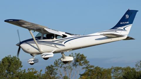 A Cessna 182 Skylane Private Aircraft, landing at the Marion Municipal Airport, Indiana. It shows a small white plane, with blue livery, in the sky, over a number of trees.
