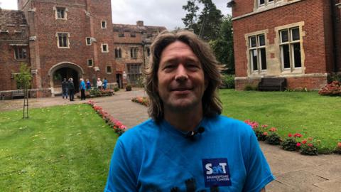 Reuben Milne wears a blue T-shirt in front of Buckden Towers, which is a red-bricked building with turrets along the top. Behind him is a grassed area with pink and red flowers. 