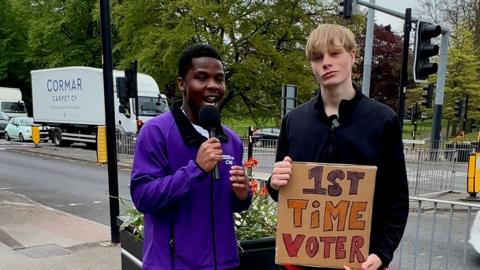 A young man in a purple jacket speaking into a microphone next to another young man in a dark top holding a cardboard sign reading 1st time voter