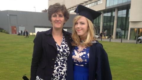 A woman with brown hair wearing a white frock and navy blazer smiles at the camera, holding her daughter, who has blonde hair and is wearing a blue dress and a graduation hat and blazer, standing outside of a university 