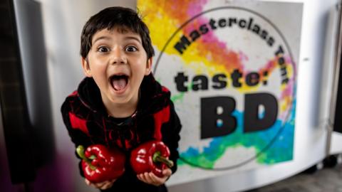 A young boy stands holding some red capsicum peppers and poses joyfully into the camera. He stands in from of a sign that says Masterclass Tent - Taste: BD