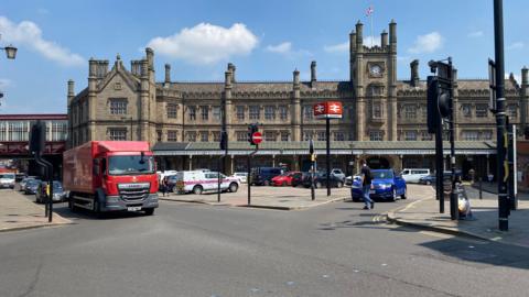 Shrewsbury railway station is in the background, it is a large brown building with a clock tower and dozens of high chimneys. There is a car park in front of it and a road running parallel to it. A red lorry is driving on the road, through some traffic lights.
