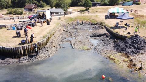 Image shows drone picture of the excavation site. The slip way is exposed but partially covered by water at the bottom. To the left of the picture, there is a diver stood near the water with diving gear on. On the grass, is the rest of the diving team with a white truck which is storing the equipment. In the middle, you can see the exposed groundways. To the right of the image, is a green and white tent which is open to the public to see the artefacts found and explaining what is taking place.