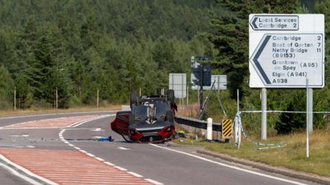 The car is on its roof. Doors on one side of the dark red vehicle are open and there is debris strewn across the carriageway of the A9 near Carrbridge. The car is next to a metal barrier and there is blue and white police tape next to a large road sign. The sign shows the direction and miles to a number of destinations, including Carrbridge, Boat of Garten and Grantown on Spey. There is a large area of forestry in the background.