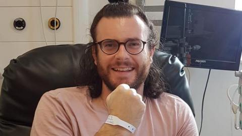 Wearing a pink t-shirt and with a hospital ID tag around his right wrist, James Carroll smiles open mouthed at the camera. He sits in a hospital chair, wearing glasses and with long, brown hair tied in a bun.