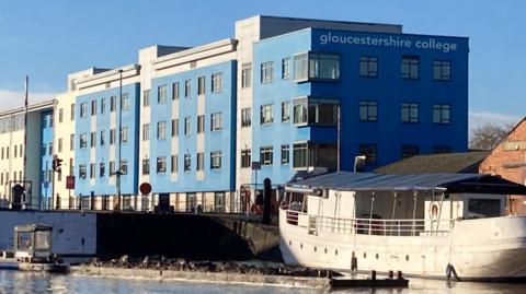 A photo of the Gloucestershire College campus next to Gloucester Docks. The building has blue and white cladding and is next to canal basin, where a white boat is moored up