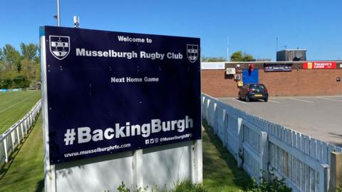 A sign for Musselburgh Rugby Club, with the club building in the background. The sky is blue. There is a black car parked outside.