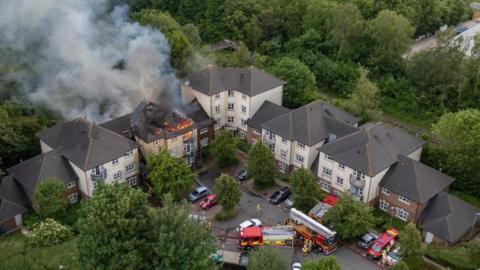 Blocks of flats in a V-shape with several fire engines on the road outside - one block is on fire with flames and smoke coming from its roof.