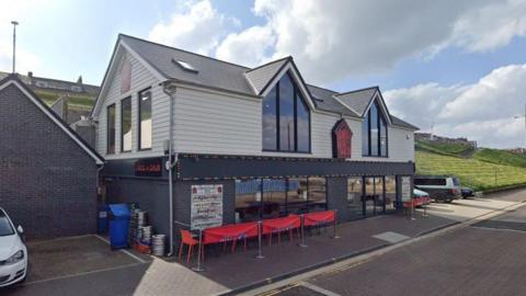 An external view of Grandpa Dickie's Shed on Marine Walk. Red chairs are placed outside the restaurant, which has large glass windows.