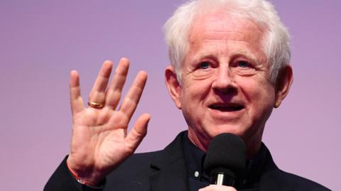 Richard Curtis waving at the camera holding a microphone. He is dressed in a black suit. 