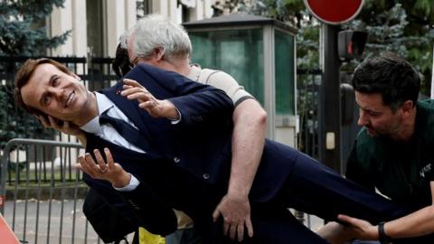 Two men carry the suited waxwork of Emmanuel Macron outside the Russian embassy in Paris