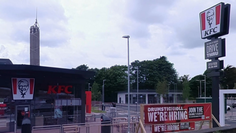 The exterior of KFC, with a drive-through sign reading KFC with firms logo, which is an image of an older man. It sits in a large car park. 