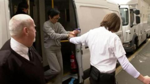 Papaipit Linse arriving at court. She is being led off a prison van by two employees, a woman wearing a white shirt and a man dressed in a black top. Papaipit is wearing an all-grey prisoned issued jogging bottoms and jumper. She is handcuffed to the woman wearing white