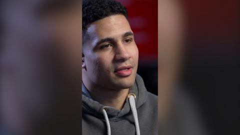 Delicious Orie looks off camera. He has short black hair and is wearing a grey hoodie with white drawstrings.