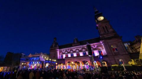 Night-time view of hundreds of people watching a performance under a canopied stage at night outside The Atkinson cultural venue, which is an old stone building with an illuminated clock tower in Southport.