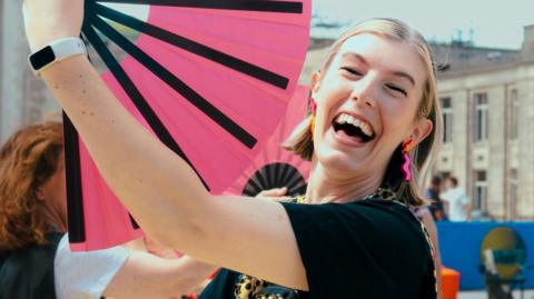 Dancers and members of the public are holding bright pink fans, with one lady with blond hair, wearing an animal print top, smiling at the camera