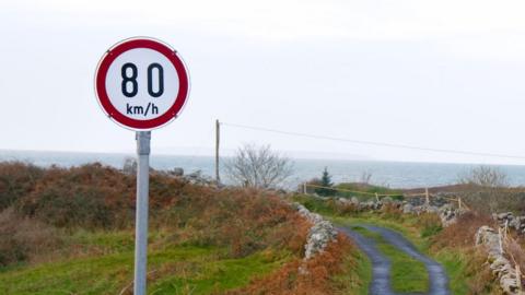 A speed limit sign on a small country road says 80km/h (50mph). The road has grass growing in the middle. The sea can be seen over the crest of the hill. 