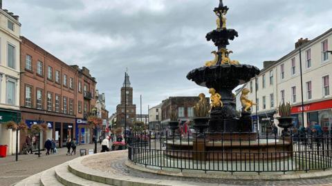 The centre of the market town of Dumfries with a fountain in the foreground and its Midsteeple building in the distance