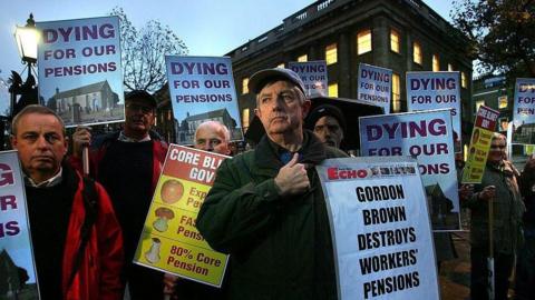 A group of men protesting in central London holding signs saying "Gordon Brown destroys workers' pensions" and "dying for our pensions". 