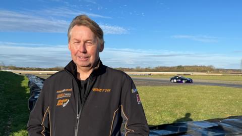 Alan Mugglestone stands at the side of a motor circuit in Lincolnshire which is bathed in bright sunshine and blue skies. He has medium length brown hair and is wearing a black jacket covered in orange logos. A black racing car is visible in the background.