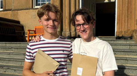 Archie and Charlie - Archie has light brown hair long on the top short on the sides, smiling holding a large brown envelope wearing a white blue and red striped shirt, Charlie is smiling with teeth same hair as Archie but brown, black rimmed round glasses, white shirt also holding a brown envelope, background of stairs and a brown building with two light brown benches