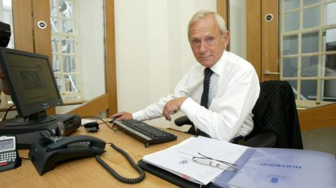 Sir George Reid in white shirt and dark tie at his desk when he was Presiding Officer in Holyrood. It is a small, modern office.