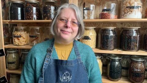 Sam is looking into the camera and smiling slightly. She has chin-length silver hair, and is wearing a pair of glasses. She's got a mustard yellow cable knit jumper on, with a teal blue woollen cardigan over the top. Sam is also wearing a blue denim apron, which has a small house logo embroidered on it in white thread. Behind her are six wooden shelves, all of which have rows of large glass jars on them.