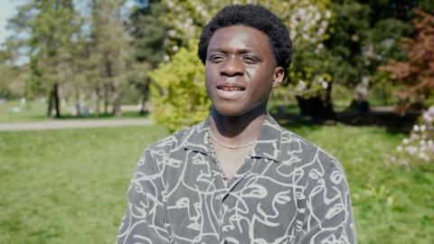 A young black manis wearing a grey-white patterned shirt. The background shows green trees and grass in a park with bright sunshine.