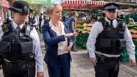 Home Secretary Yvette Cooper walks past market stalls in Lewisham town centre, London, with two police officers in uniform in July 2024.