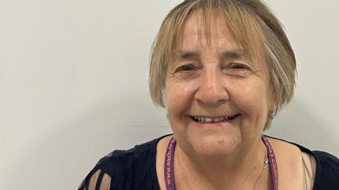 Hazel Brown has short blonde and light brown hair. She is standing in front of a white wall and is wearing a black T-shirt and a purple lanyard.