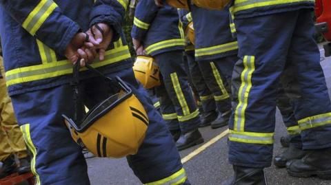 A view of the legs and lower bodies of a group of several firefighters in navy uniform with yellow fluorescent stripes. They are holding yellow helmets.