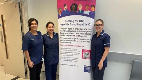 Three nurses in dark blue unifroms are stood in front of a white wall in a corridor , In the middle of them is a white display board with a cartoon of peop,e at the top and writing below